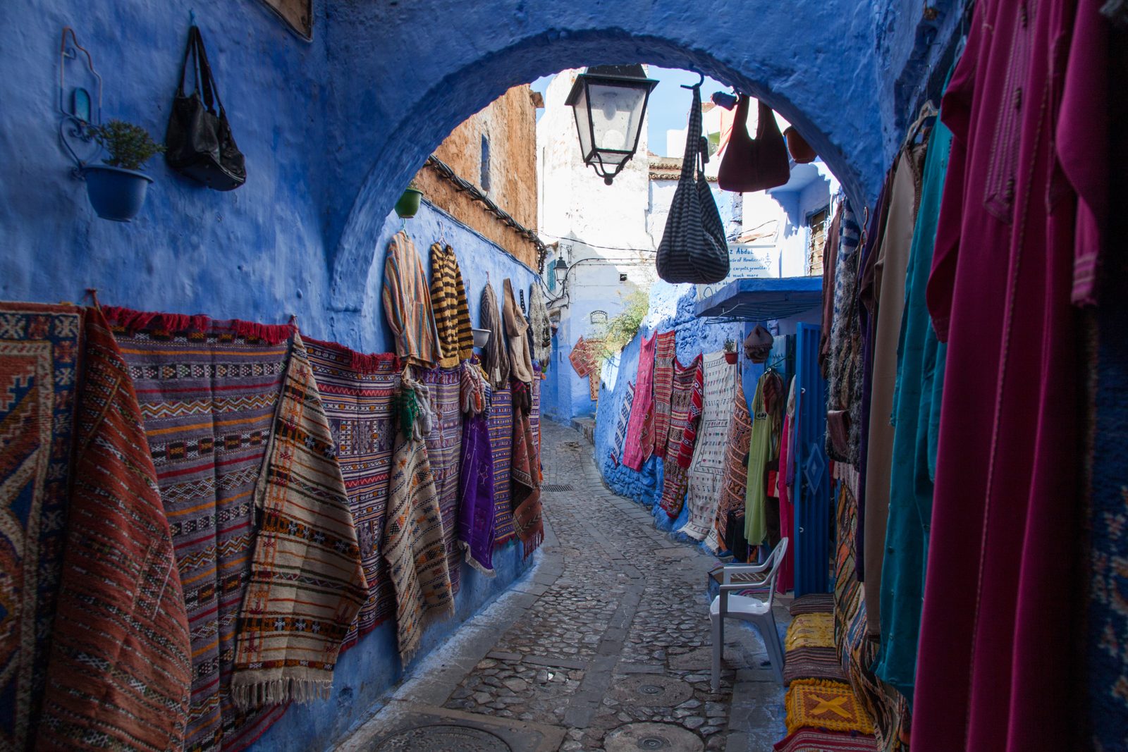 A blue-painted alley with woven rugs and kaftans hanging under an arch